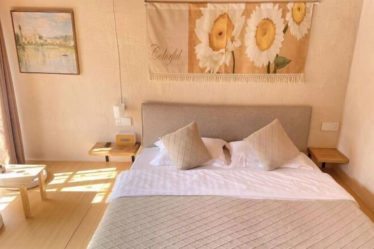 A neatly made bed with a quilted cover, two pillows, and a headboard. There is a small bedside table with a lamp and a framed picture on the wall.