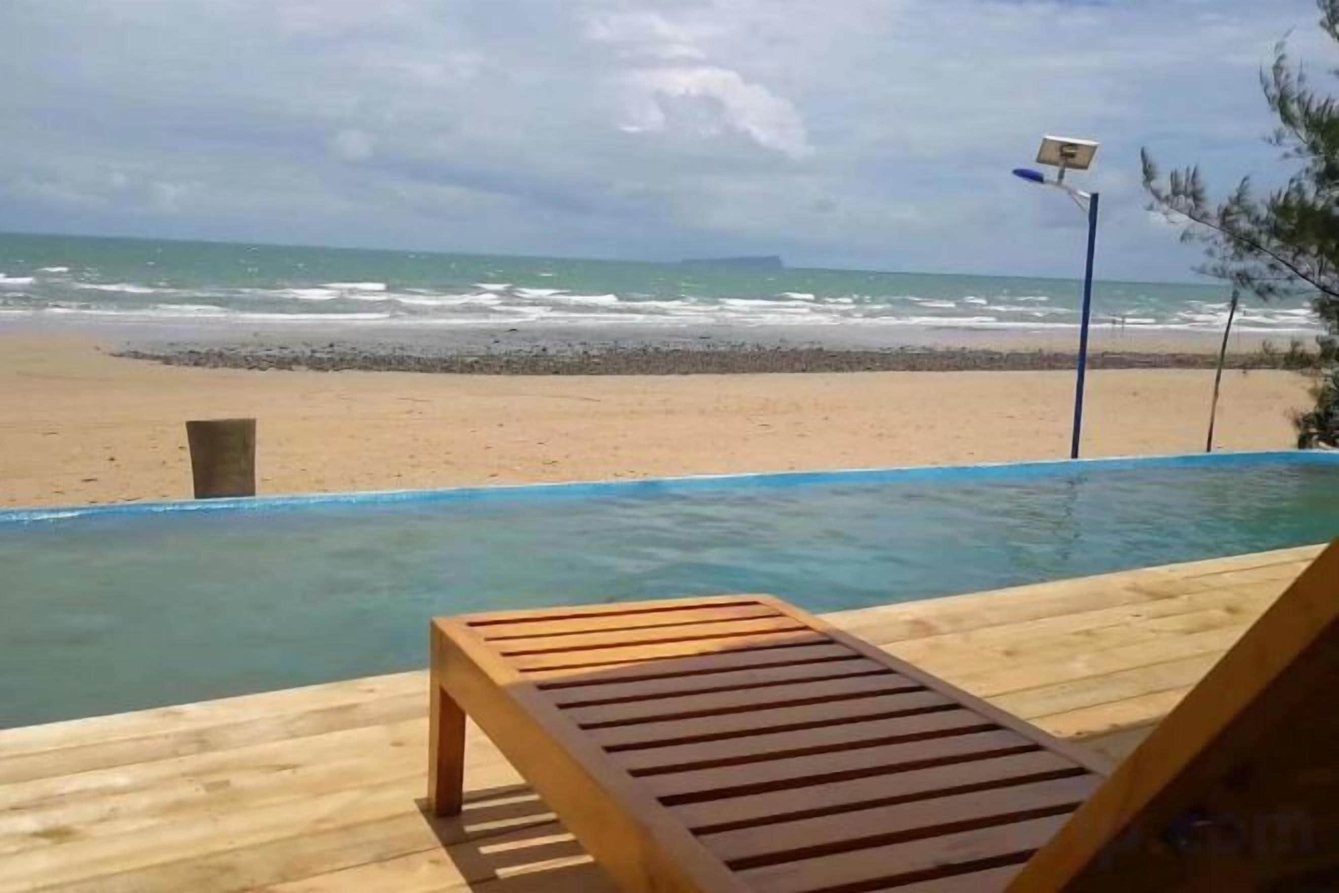 Una piscina infinita con cubierta de madera y una silla de descanso, con vista a una playa con olas y cielo nublado.