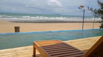 Una piscina infinita con cubierta de madera y una silla de descanso, con vista a una playa con olas y cielo nublado.