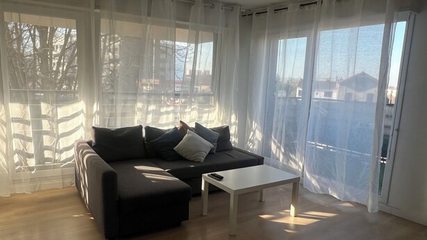Un salon moderne avec un canapé de couleur foncée, une table basse blanche, des rideaux légers et une vue sur un quartier résidentiel.