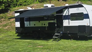A Forest River RV with a black awning, a ramp, and a logo on the side. - New River RV Travel Trailer with 1 King bedroom and bunks - 2 small children. (Prince)