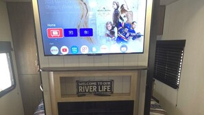A compact living space with a mounted TV displaying a sports event, a fireplace, and a sign reading "Welcome to Our River Life". - New River RV Travel Trailer with 1 King bedroom and bunks - 2 small children. (Prince)