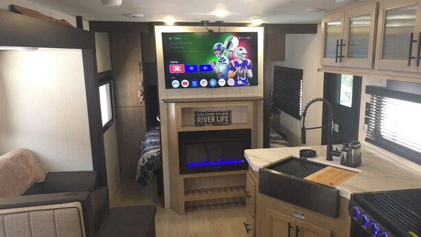 A compact RV interior with a flat-screen TV mounted on the wall, a wooden cabinet below, and a kitchenette area with a stove and sink. - New River RV Travel Trailer with 1 King bedroom and bunks - 2 small children. (Prince)
