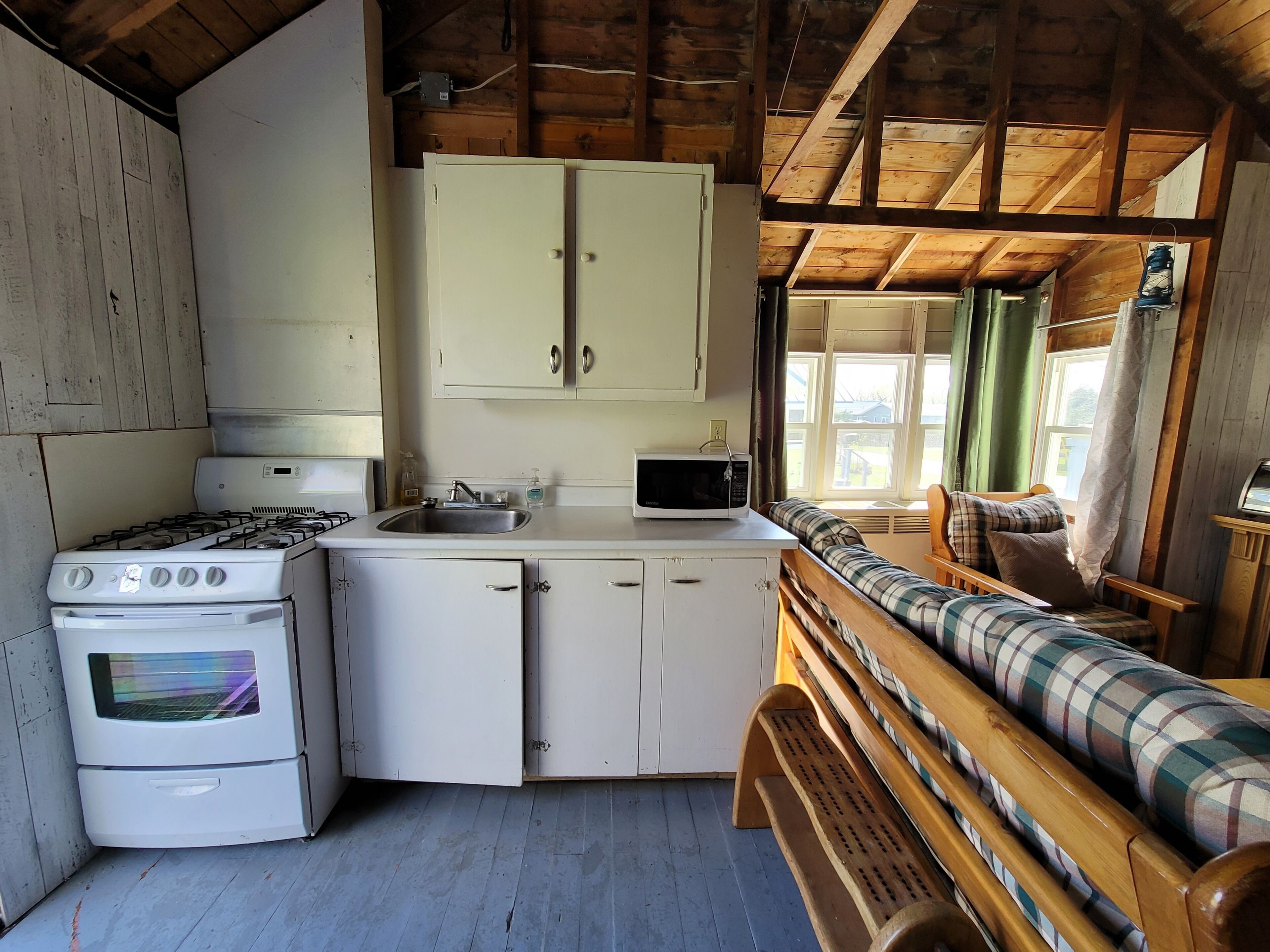 A compact kitchen with white cabinets, a stove, and a microwave. There's a bench with plaid cushions and a window with curtains.