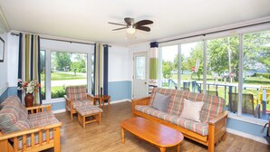 A sunlit room with a sofa, chairs, and a table. Large windows offer a view of a green landscape.