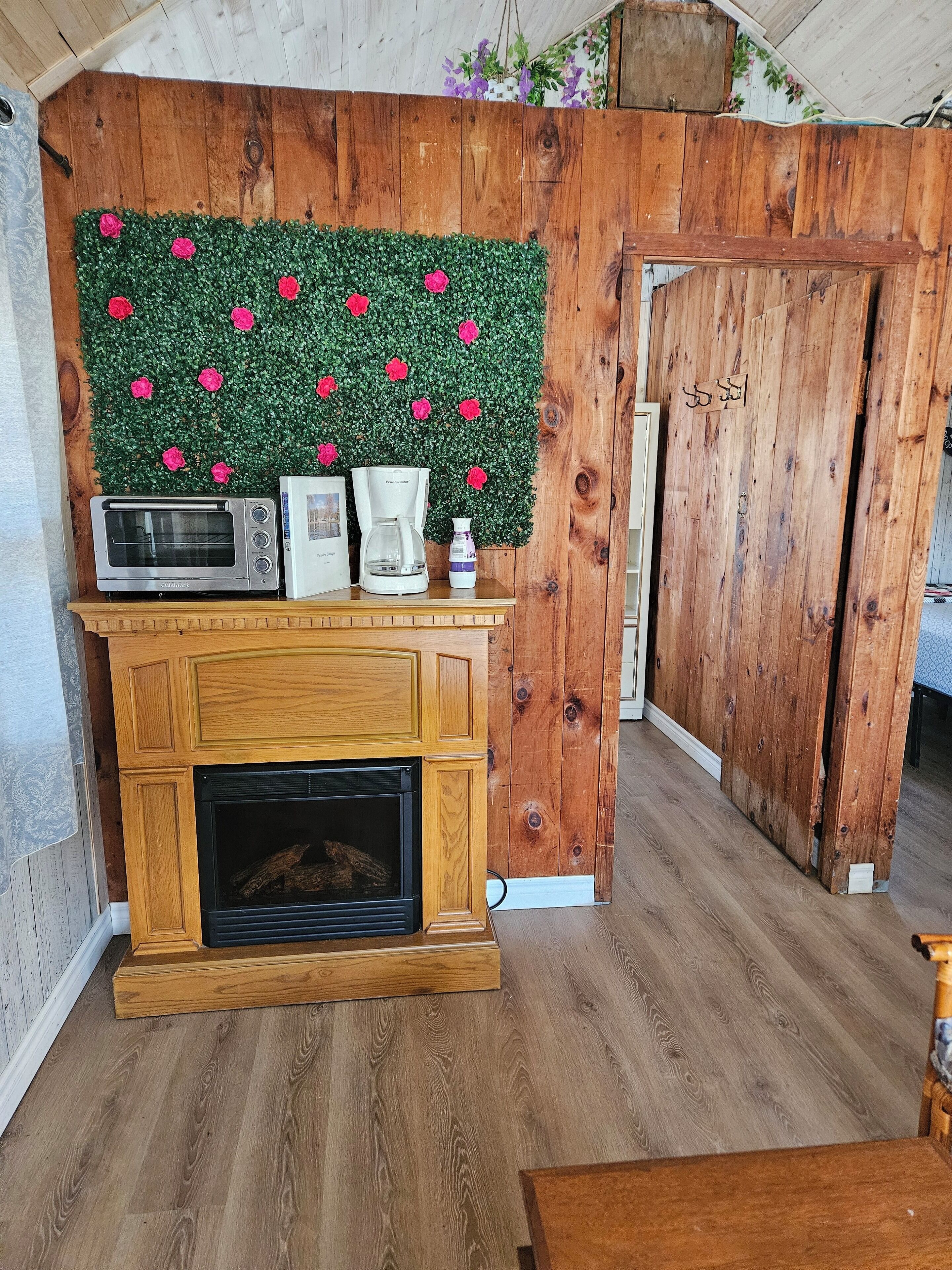 A rustic room with a wooden wall, a fireplace, a microwave, a coffee maker, and a decorative wall with pink flowers.