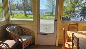 A cozy room with a wooden floor, a wicker chair, a wooden table, and large windows offering a view of a park and a car parked outside.