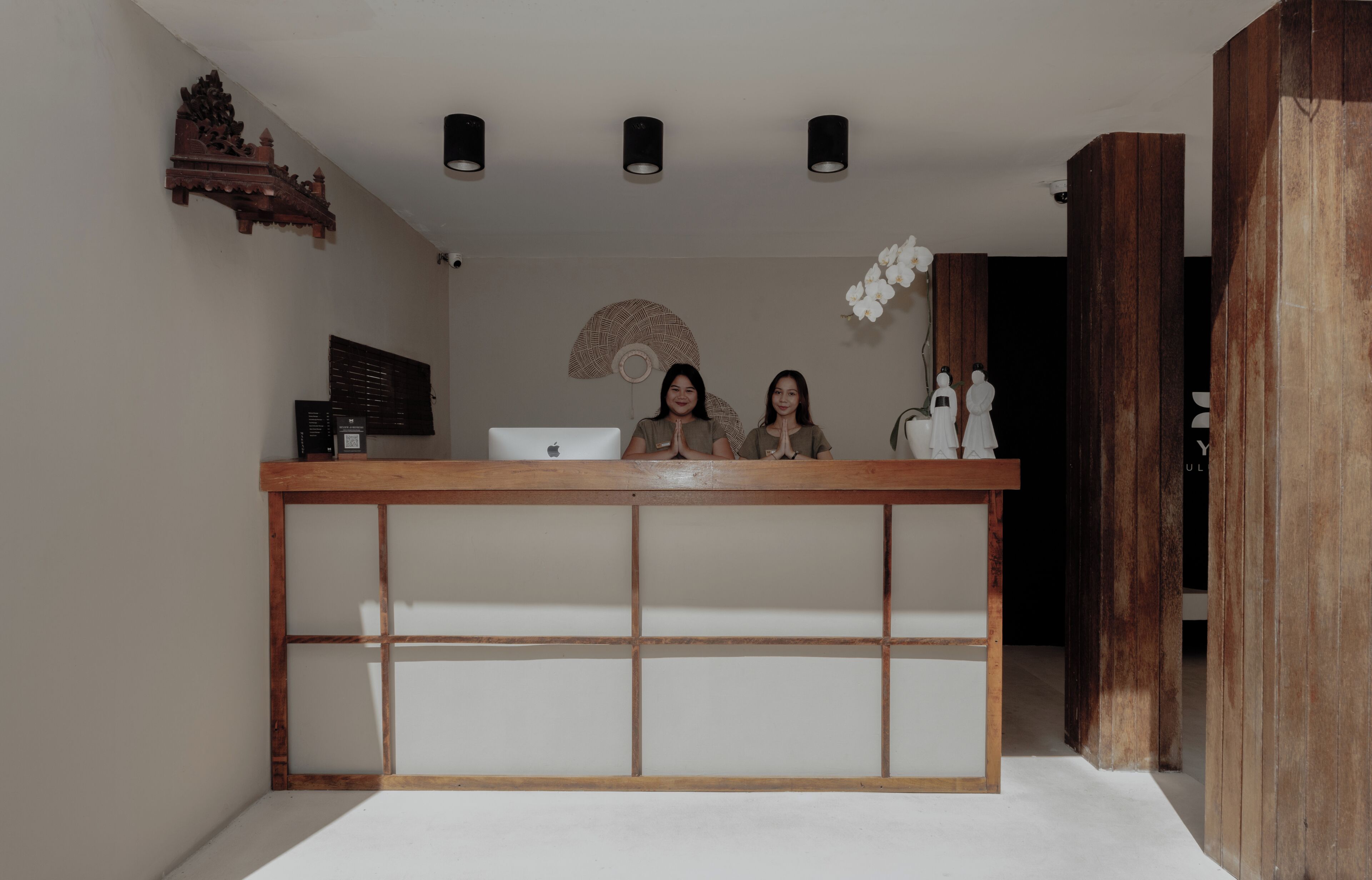Una recepción con dos personas detrás, diseño de paneles de madera y un ventilador decorativo en la pared.