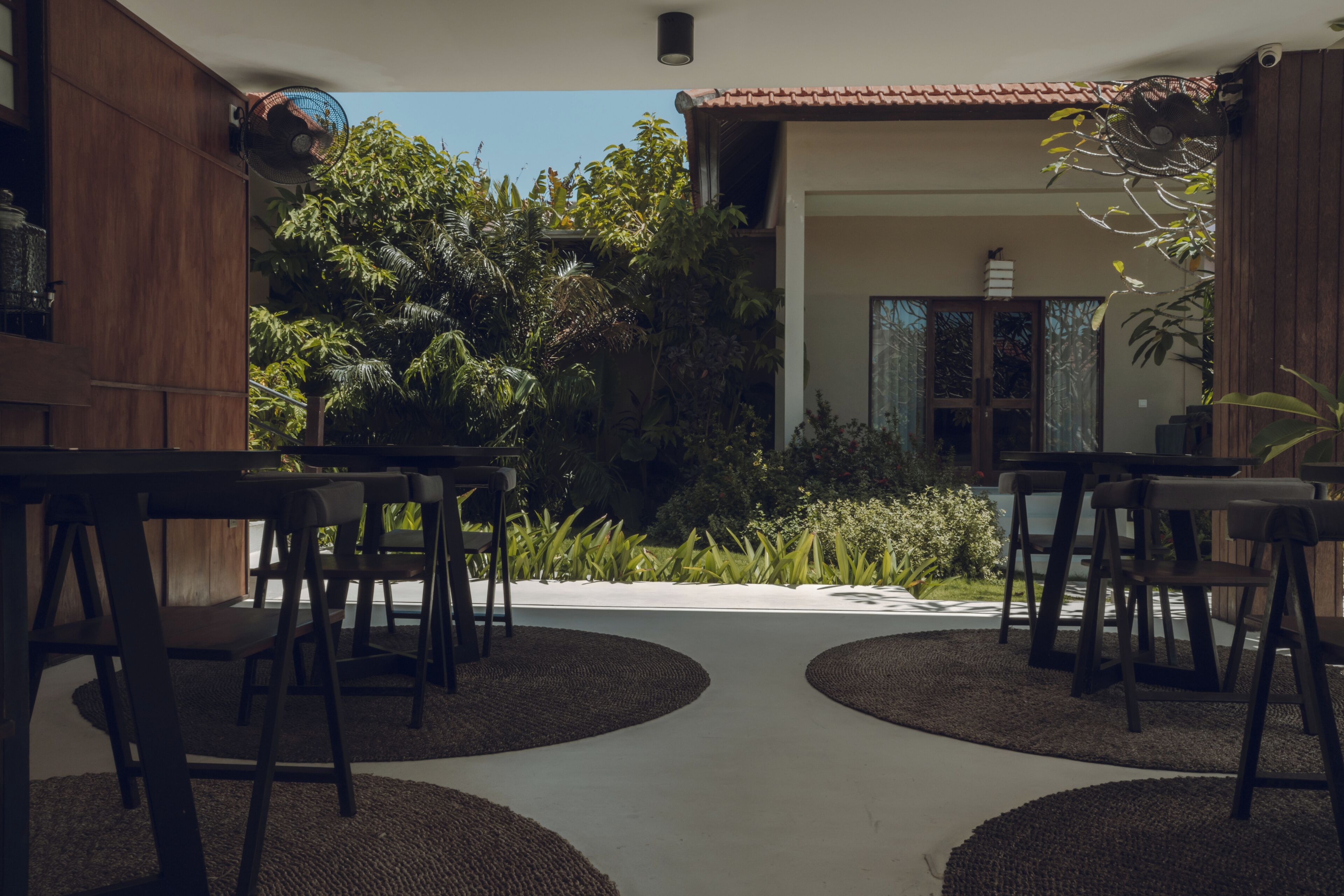 Uma área externa com mesas e cadeiras, cercada por plantas e vista para um edifício.