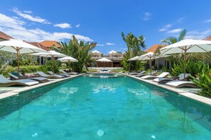 Piscina com espreguiçadeiras e guarda-sóis, rodeada por vegetação e edifícios com telhados de telha vermelha.