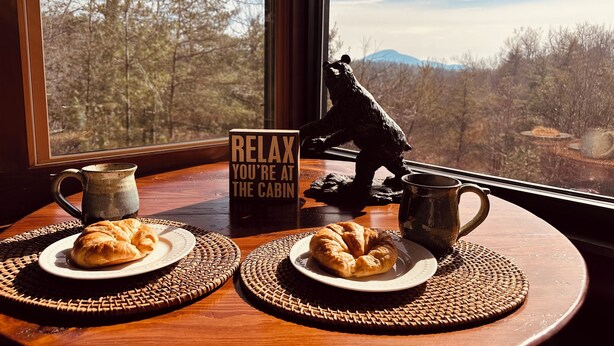 Ein Holztisch mit zwei Tellern Croissants, einer Keramiktasse und einer Bärenfigur. Ein Schild mit der Aufschrift 'RELAX YOU'RE AT THE CABIN'.