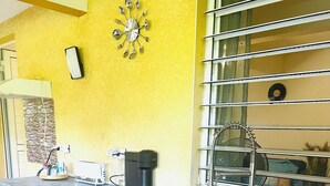 A kitchen with a yellow wall, a clock, a coffee maker, and a window with a metal frame. - Studio in a green setting 15 min from the Jarry industrial zone and 2 min from the rivers. (Petit-Bourg)