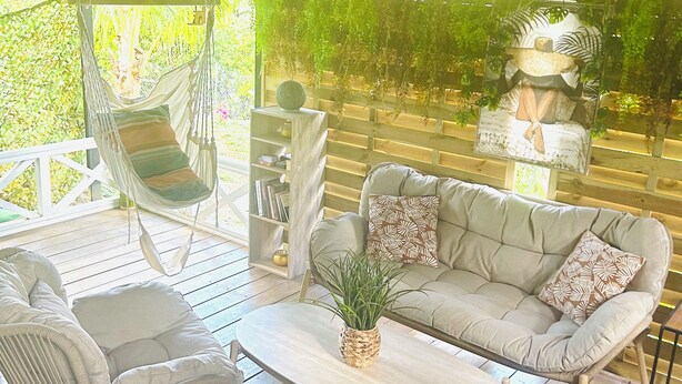 A covered outdoor seating area with wicker furniture, a wooden table, and a hanging chair. - Studio in a green setting 15 min from the Jarry industrial zone and 2 min from the rivers. (Petit-Bourg)