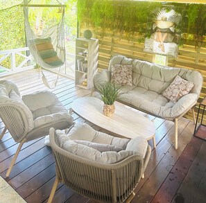 A covered outdoor seating area with wicker furniture, a wooden table, and a hanging chair. - Studio in a green setting 15 min from the Jarry industrial zone and 2 min from the rivers. (Petit-Bourg)