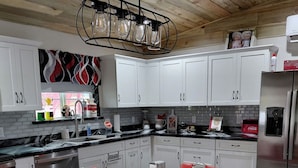 A modern kitchen with white cabinets, a stainless steel refrigerator, a round dining table with red chairs, and a unique chandelier. - Relaxing 2-bedroom cabin decorated with Coke Cola Theme, (Payson)