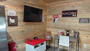 A cozy room with wooden walls, a television, a popcorn sign, a Coca-Cola refrigerator, and a small seating area with a red cushion. - Relaxing 2-bedroom cabin decorated with Coke Cola Theme, (Payson)