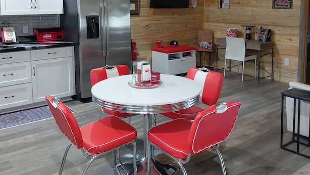 A modern kitchen with white cabinets, a stainless steel refrigerator, and a dining area with a round table and red chairs. - Relaxing 2-bedroom cabin decorated with Coke Cola Theme, (Payson)