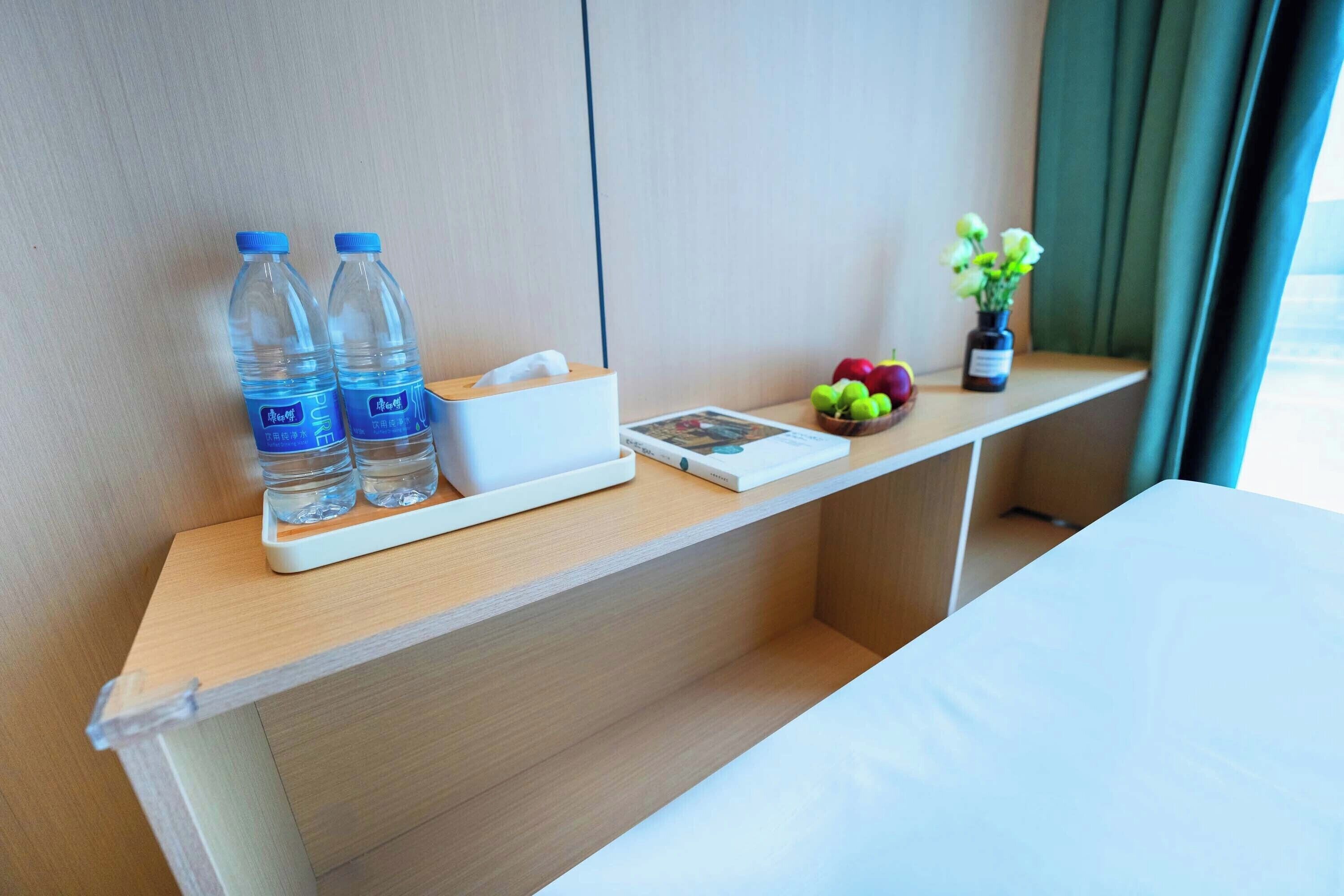 Ein Hotelzimmer mit einem Holztisch, zwei Wasserflaschen, einer Packung Taschentücher, einer kleinen Obstschale und einer Vase mit Blumen.