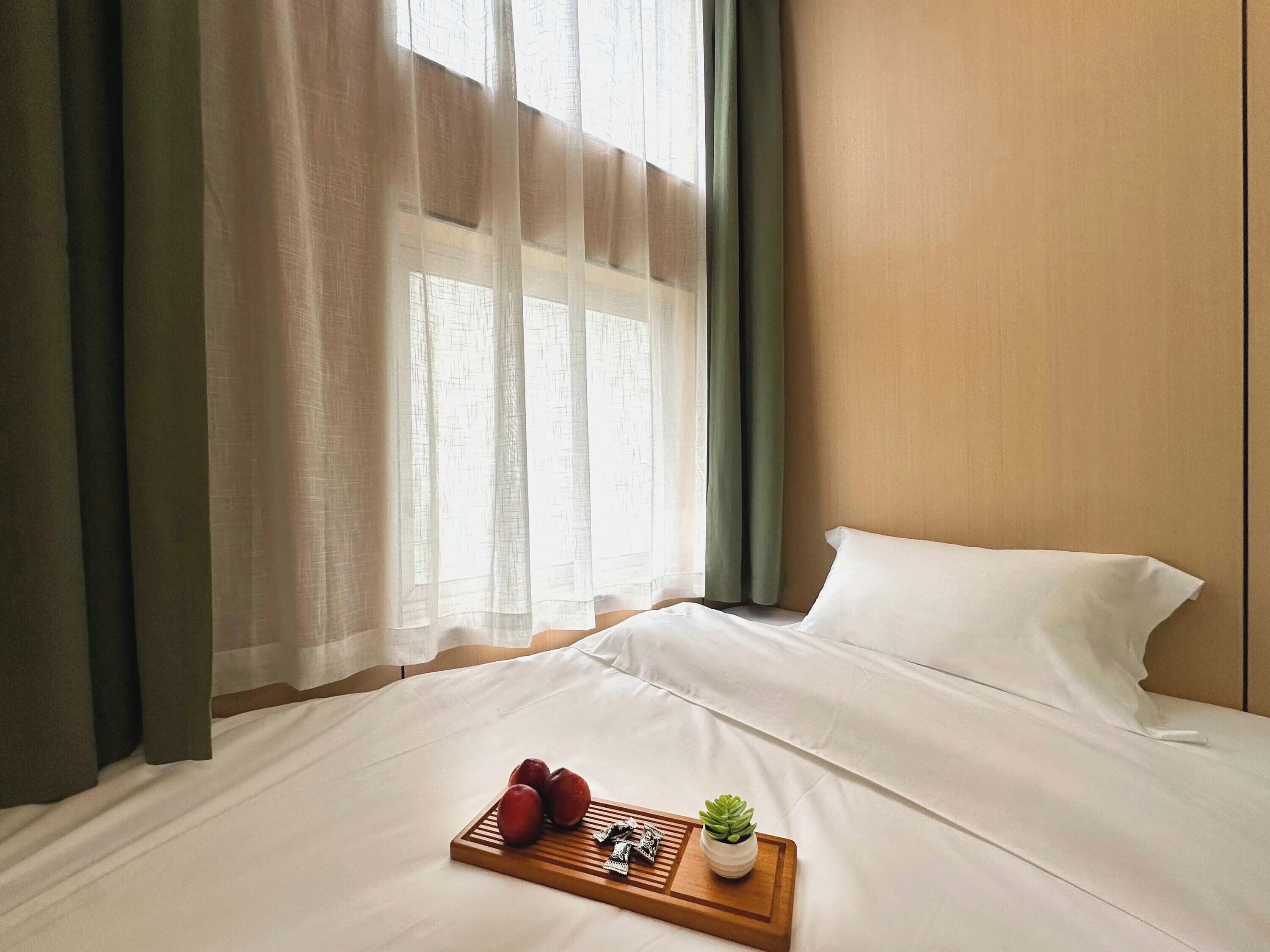 A hotel room with a bed, a tray with fruits and a small plant, and sheer curtains.