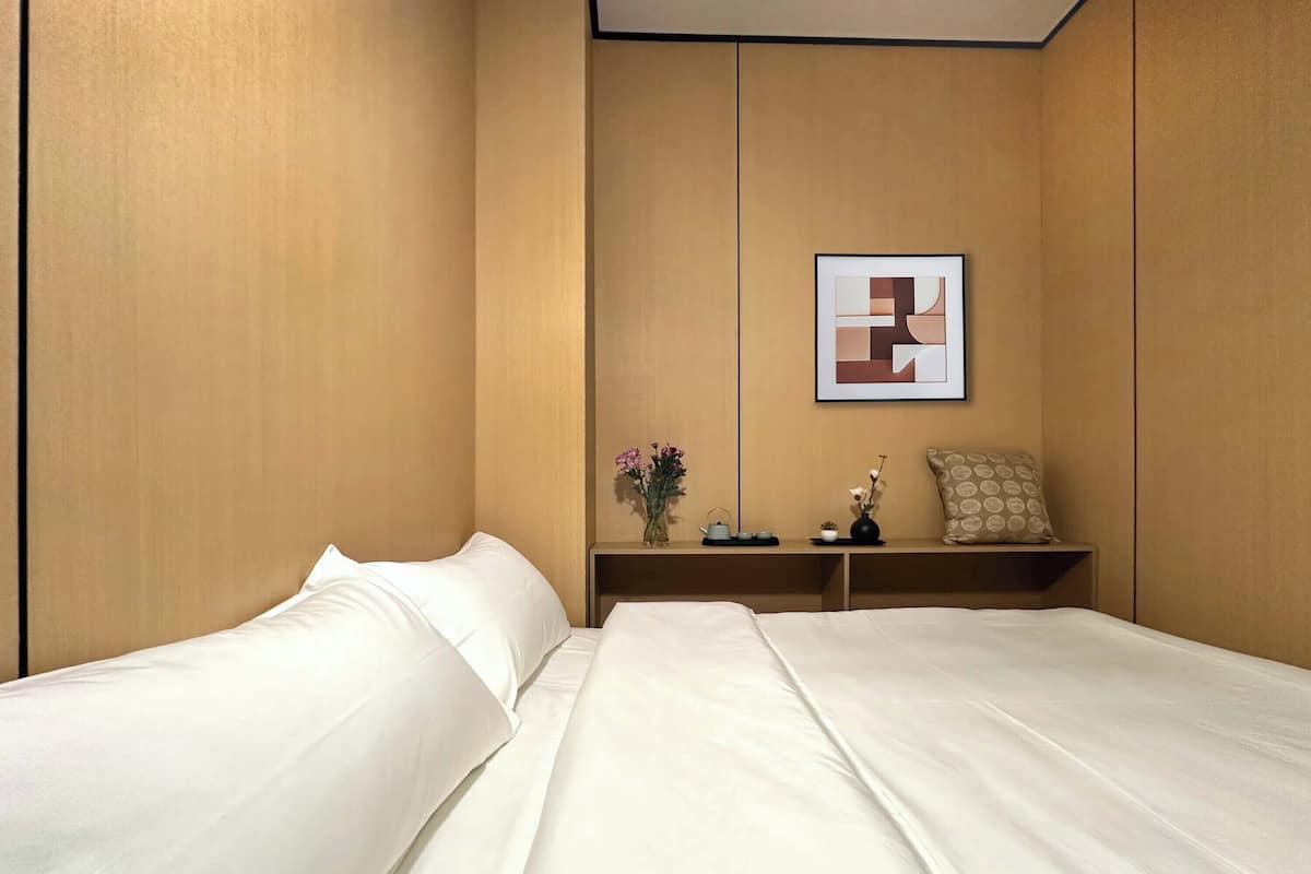 A hotel room with two beds, a nightstand, a vase with flowers, and a framed abstract artwork on the wall.