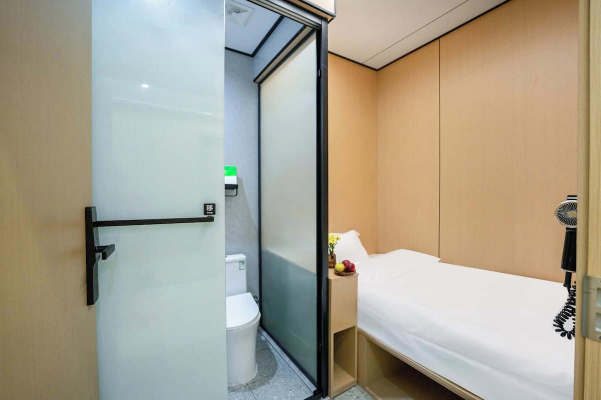 A compact hotel room with a sliding door separating the bathroom and bedroom. The bathroom features a toilet and a small sink. The bedroom has a single bed with a white comforter and a bedside table with a lamp and a small vase with flowers.