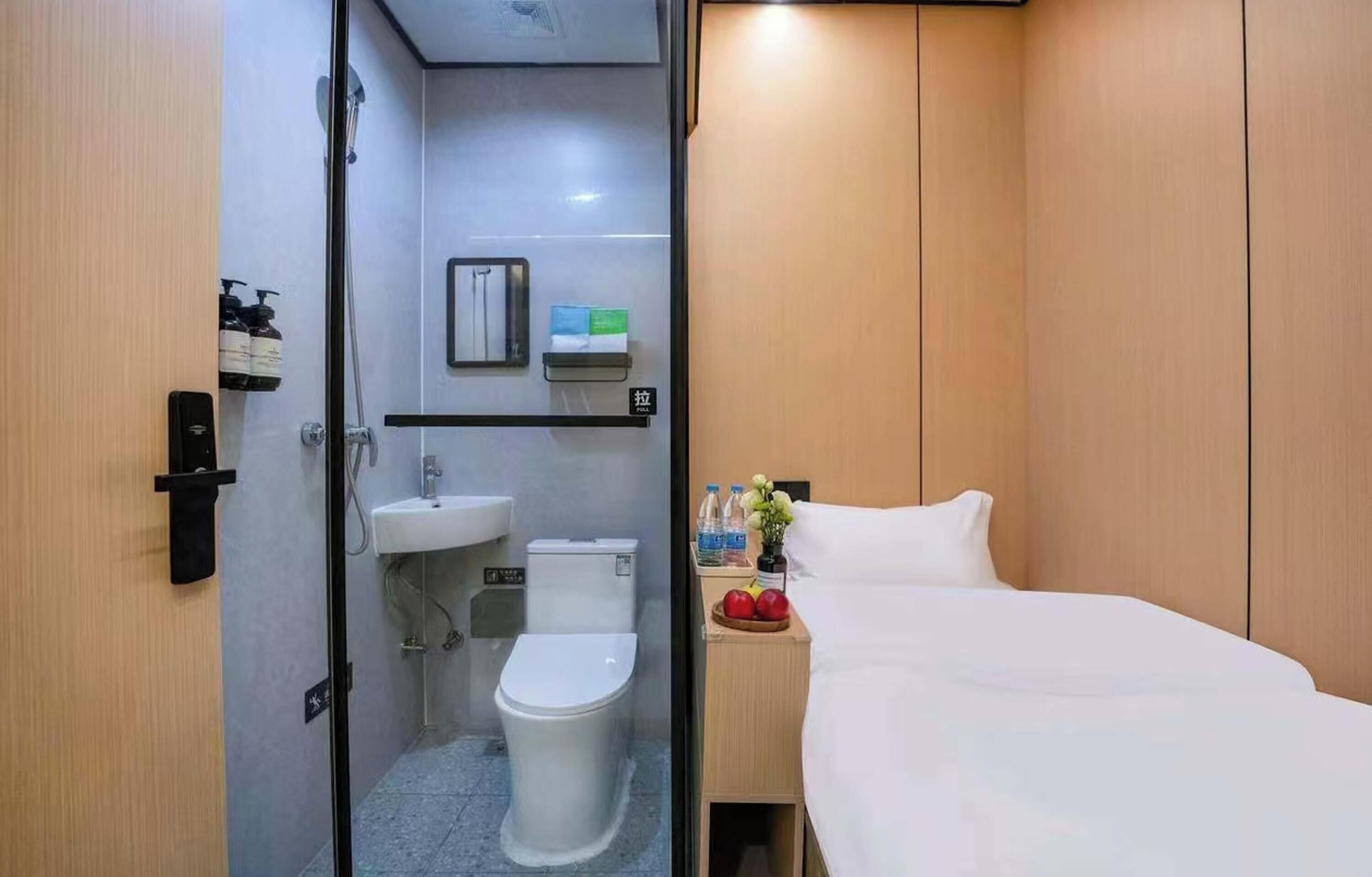 Ein Hotelzimmer mit Duschkabine, Waschbecken, Toilette, einem Bett und einem kleinen Tisch mit Früchten und einer Vase.