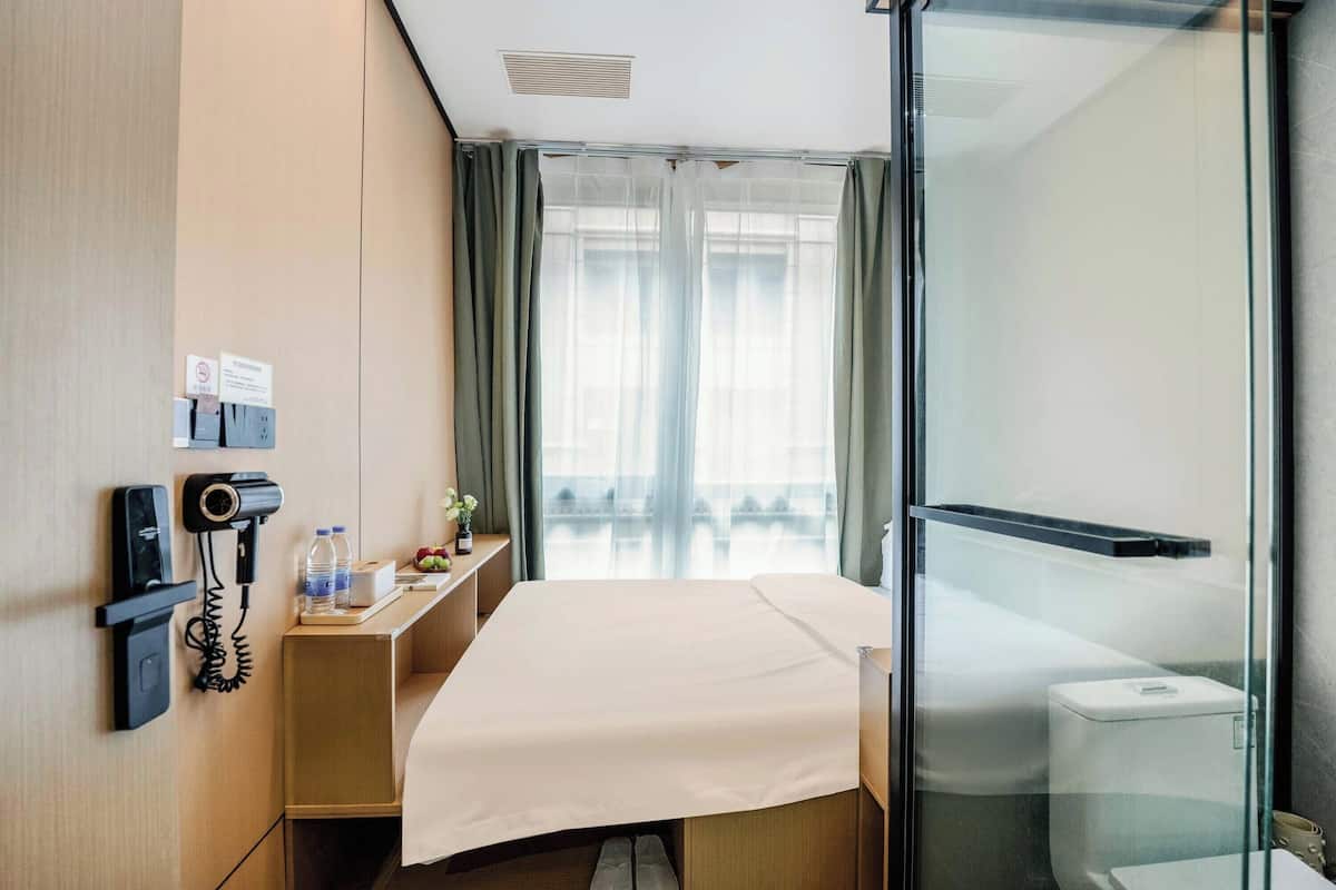 A hotel room with a bed, a desk with a phone and a lamp, a window with curtains, and a glass shower enclosure.