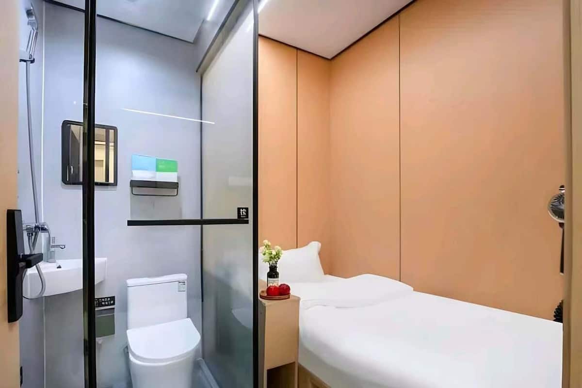 A modern hotel room with a glass-enclosed shower, a white toilet, a bed with white bedding, and a small bedside table with a vase of flowers.