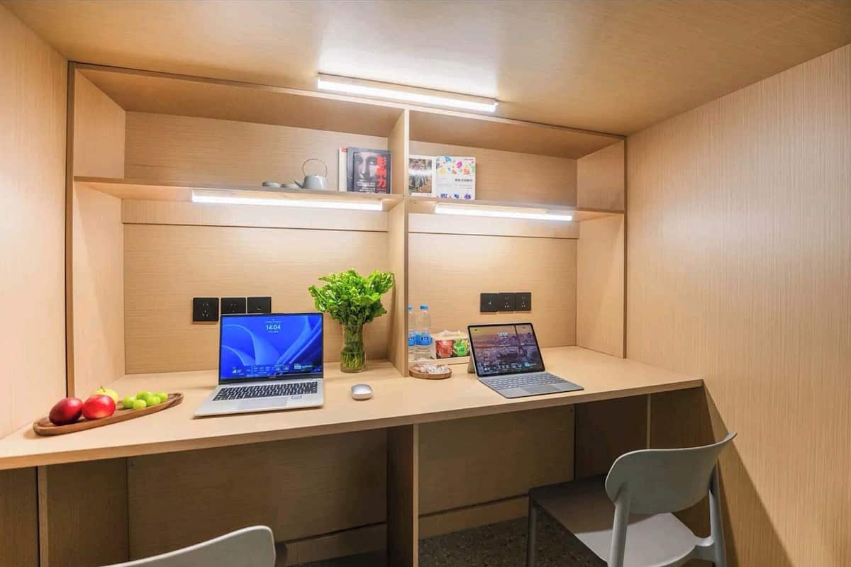 A modern office cubicle with two desks, each with a laptop, a plant, and personal items.