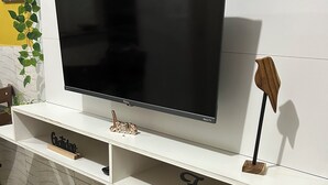Un salon moderne avec un téléviseur à écran plat posé sur un meuble blanc, des poufs à motifs floraux et une petite étagère en bois ornée d’objets décoratifs.