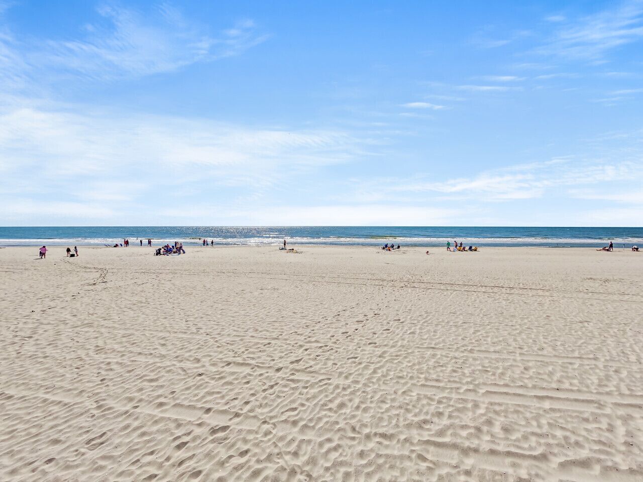 A wide sandy beach with people scattered across it, a clear sky, and the ocean in the background.