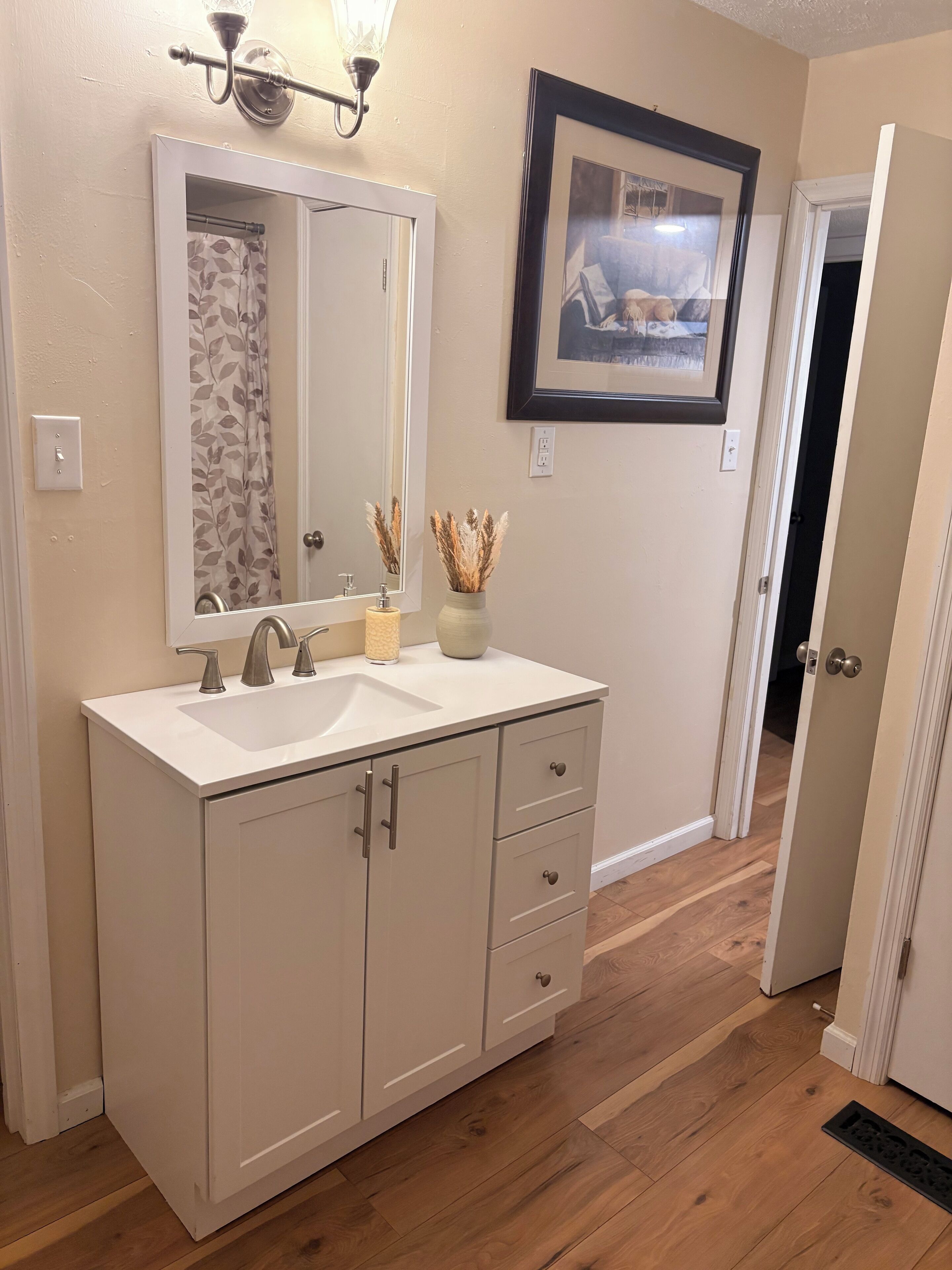 A bathroom with a white vanity, a large mirror, a shower curtain, and a framed picture on the wall.