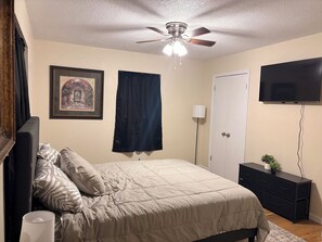 Un dormitorio con cama, ventilador de techo, televisor, cómoda y un cuadro en la pared.