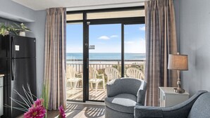 A room with a large sliding glass door leading to a balcony with beach views. There is a black refrigerator, a gray armchair, and a small table with a lamp. - Oceanfront Condo with Heated Pools (North Myrtle Beach)