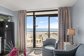 A room with a large sliding glass door leading to a balcony with beach views. There is a black refrigerator, a gray armchair, and a small table with a lamp. - Oceanfront Condo with Heated Pools (North Myrtle Beach)