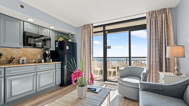 A modern kitchen with grey cabinets, a black refrigerator, and a dining area with a grey sofa and a table. - Oceanfront Condo with Heated Pools (North Myrtle Beach)