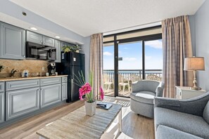 A modern kitchen with grey cabinets, a black refrigerator, and a dining area with a grey sofa and a table. - Oceanfront Condo with Heated Pools (North Myrtle Beach)