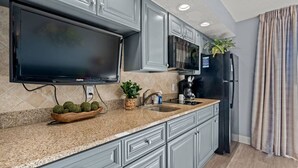 A modern kitchen with grey cabinets, a flat-screen TV, a coffee maker, and a refrigerator. - Oceanfront Condo with Heated Pools (North Myrtle Beach)