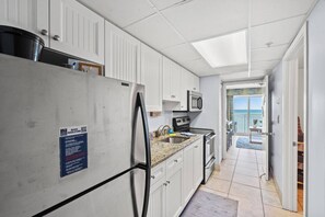 Eine Küche mit weißen Schränken, einem Edelstahl-Kühlschrank, einer Mikrowelle und einem Herd. Auf dem Kühlschrank befindet sich ein Schild und durch eine offene Tür ist der Strand zu sehen.