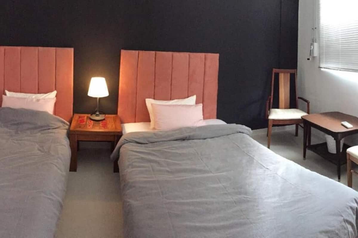 A hotel room with two single beds, a nightstand with a lamp, a small table with a chair, and a window with blinds.