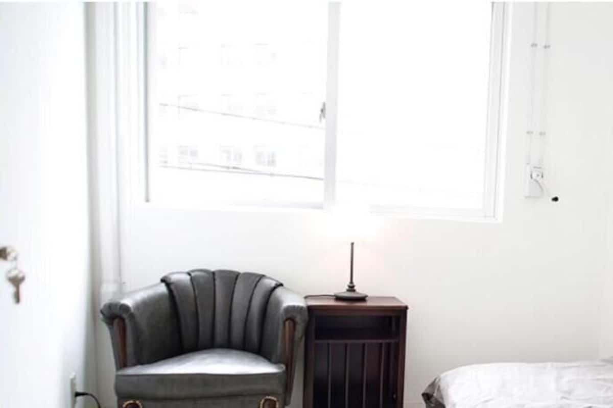 A room with a single bed, a chair, a small table with a lamp, and a window with blinds.