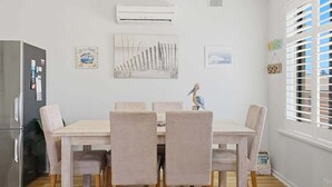 A dining area with a wooden table and chairs, an air conditioning unit, and a window with blinds. - Belle Escapes - Life is a Beach (Holdfast Bay)