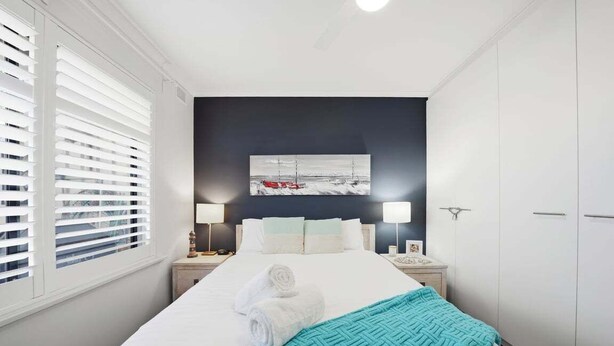 A hotel room with a large bed, white walls, a dark accent wall with a painting, and a window with blinds. - Belle Escapes - Life is a Beach (Holdfast Bay)