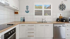 A modern kitchen with white cabinetry, a stainless steel dishwasher, a black stove, and a window with white shutters. - Belle Escapes - Life is a Beach (Holdfast Bay)