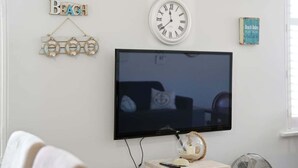 A living room with a flat-screen TV mounted on the wall, a clock, and a small shelf with decorative items. - Belle Escapes - Life is a Beach (Holdfast Bay)