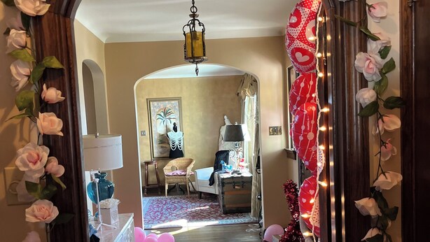 En hall med en spegelbåge dekorerad med rosa rosor, som leder in till ett sovrum med en säng, byrå och en lampa.