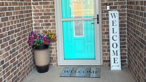A turquoise door with a white welcome sign, a potted plant with colorful flowers, and a doormat.