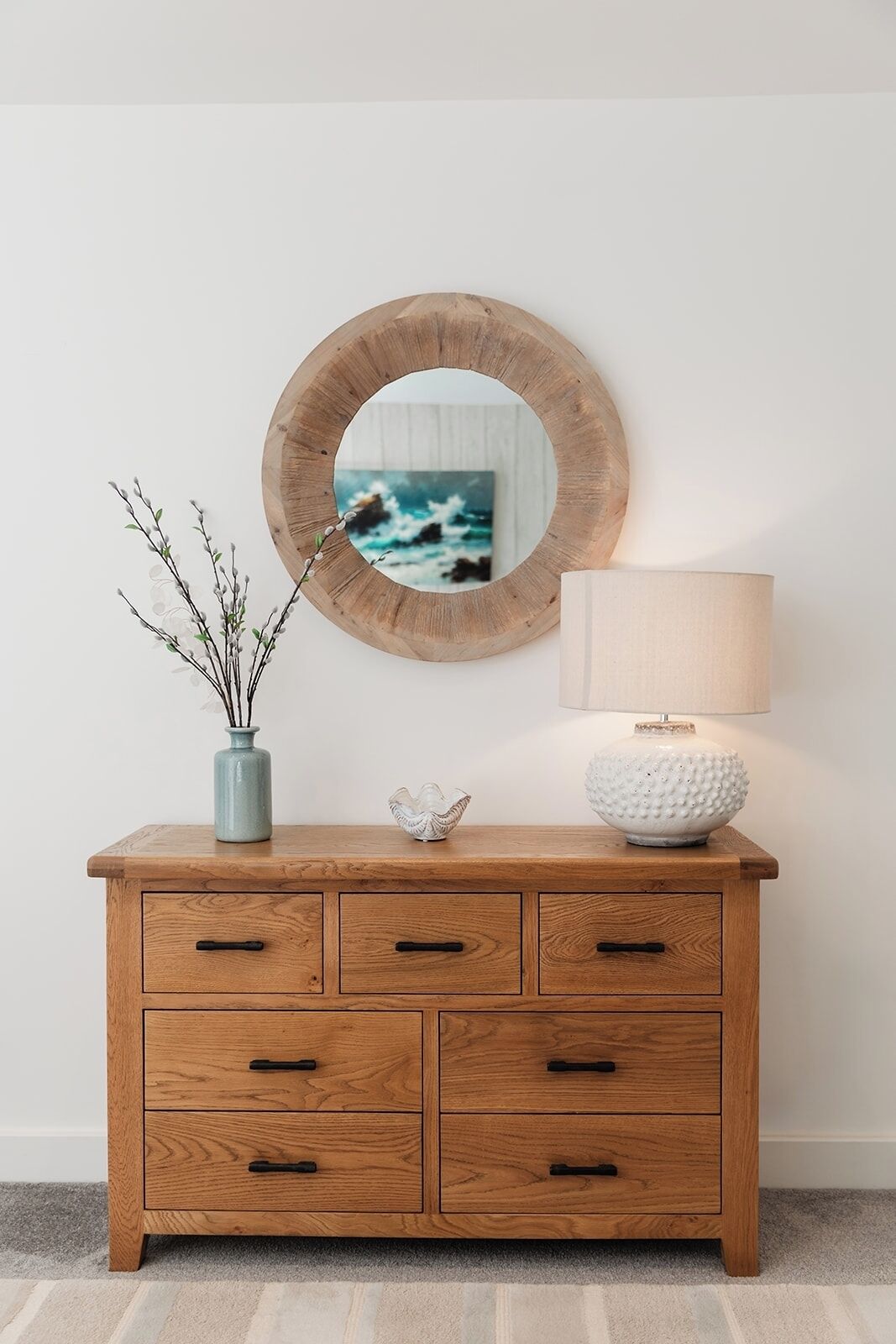 Une commode en bois avec trois tiroirs, un miroir rond, un vase avec des branches, un bol blanc et une lampe.