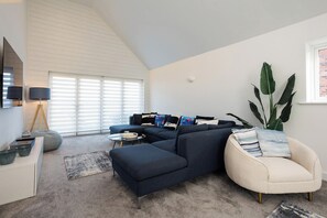 Un salon moderne avec un canapé d’angle, une table basse ronde et une petite table d’appoint. On y trouve une grande fenêtre avec des stores, un lampadaire et une plante en pot.