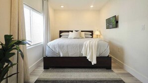A bedroom with a bed, a bedside lamp, a wall-mounted TV, and a window with curtains. - Cozy Downtown Dallas Urban Glow Retreat (Dallas)